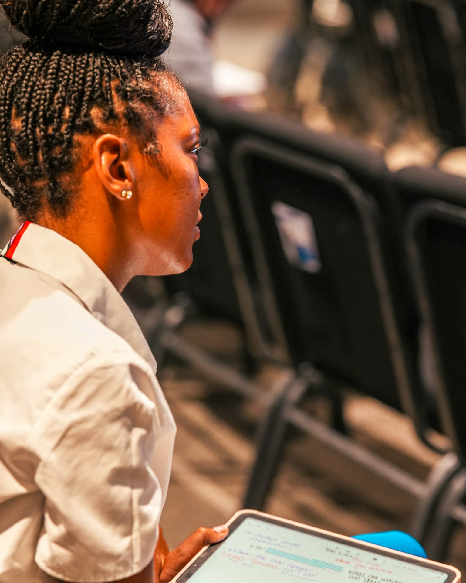 A woman in a white shirt holding a tablet while seated among rows of chairs.