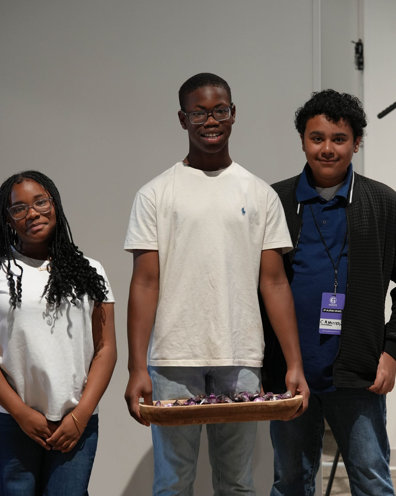 Three teenagers standing indoors while one holds a wooden tray of wrapped candies.