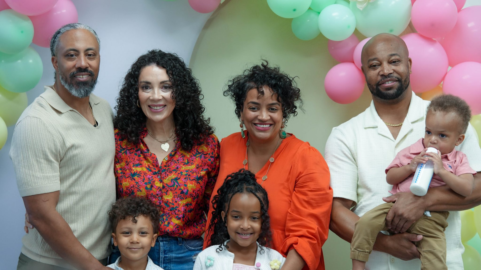 Four adults and three children standing together indoors beneath pink and green balloons, with one adult holding a toddler.