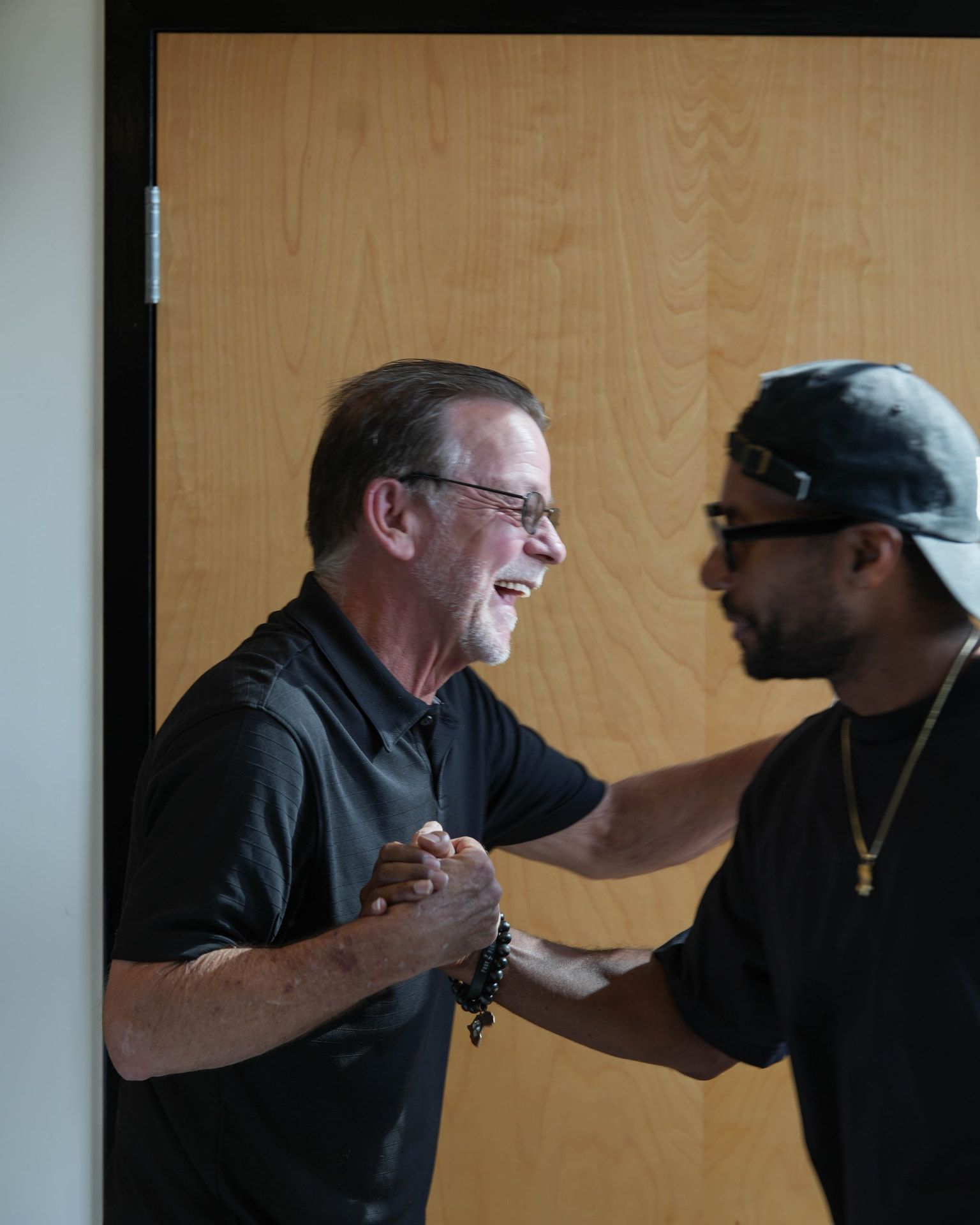 Two men smiling and clasping hands in an indoor doorway.