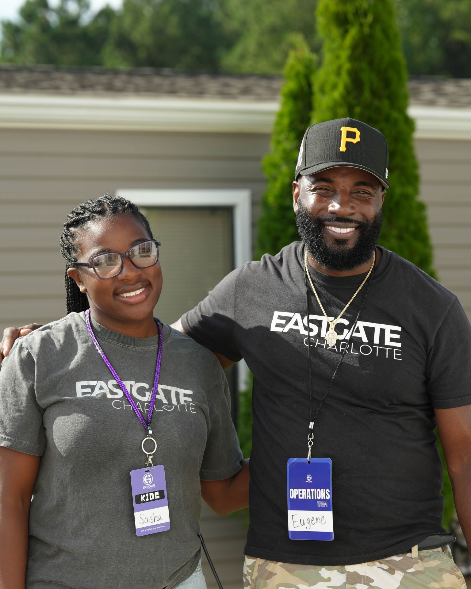 A man in a black cap smiling with his arm around a young woman in a matching t-shirt outside a building.