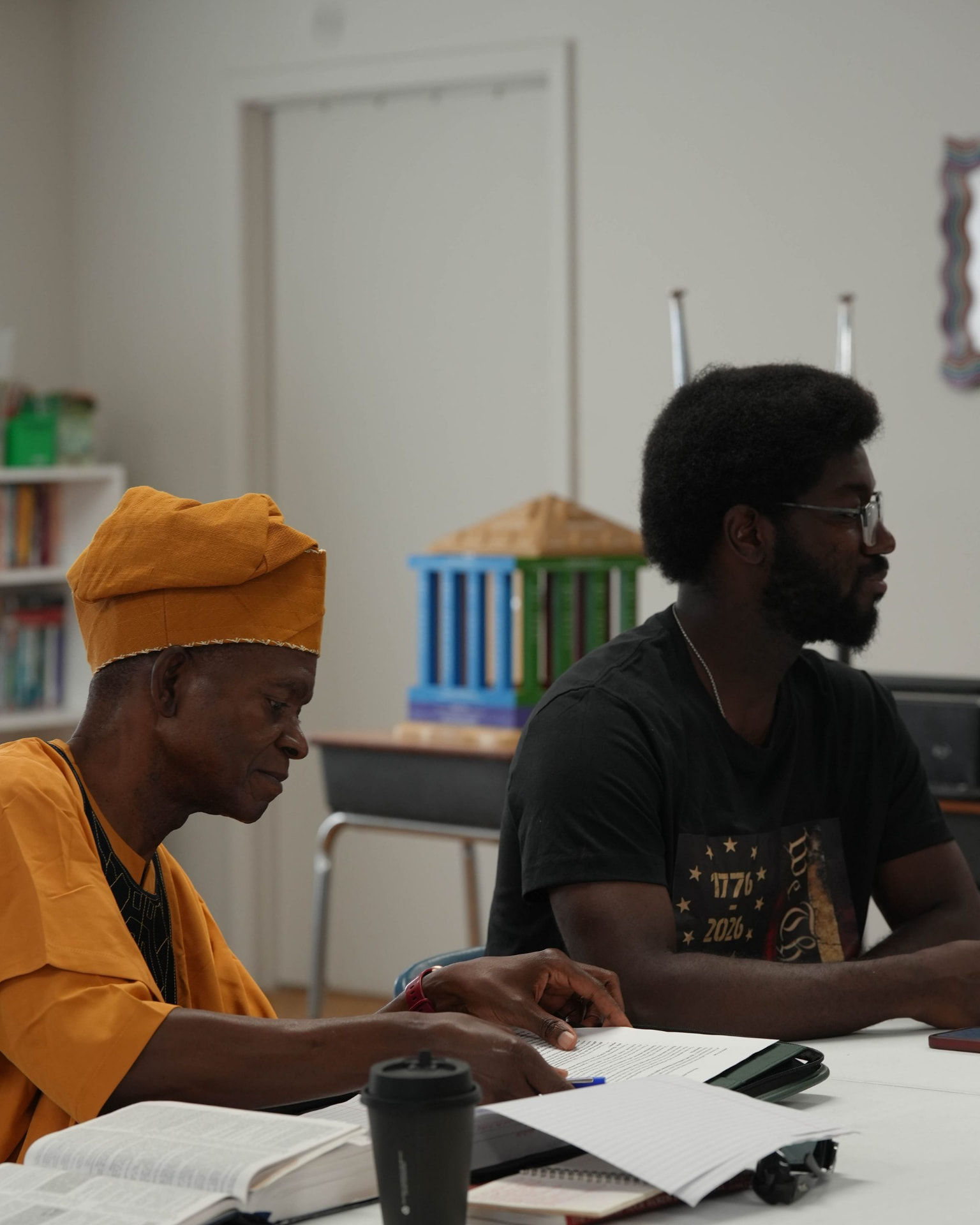 Two adults seated at a classroom table with open books and printed papers.