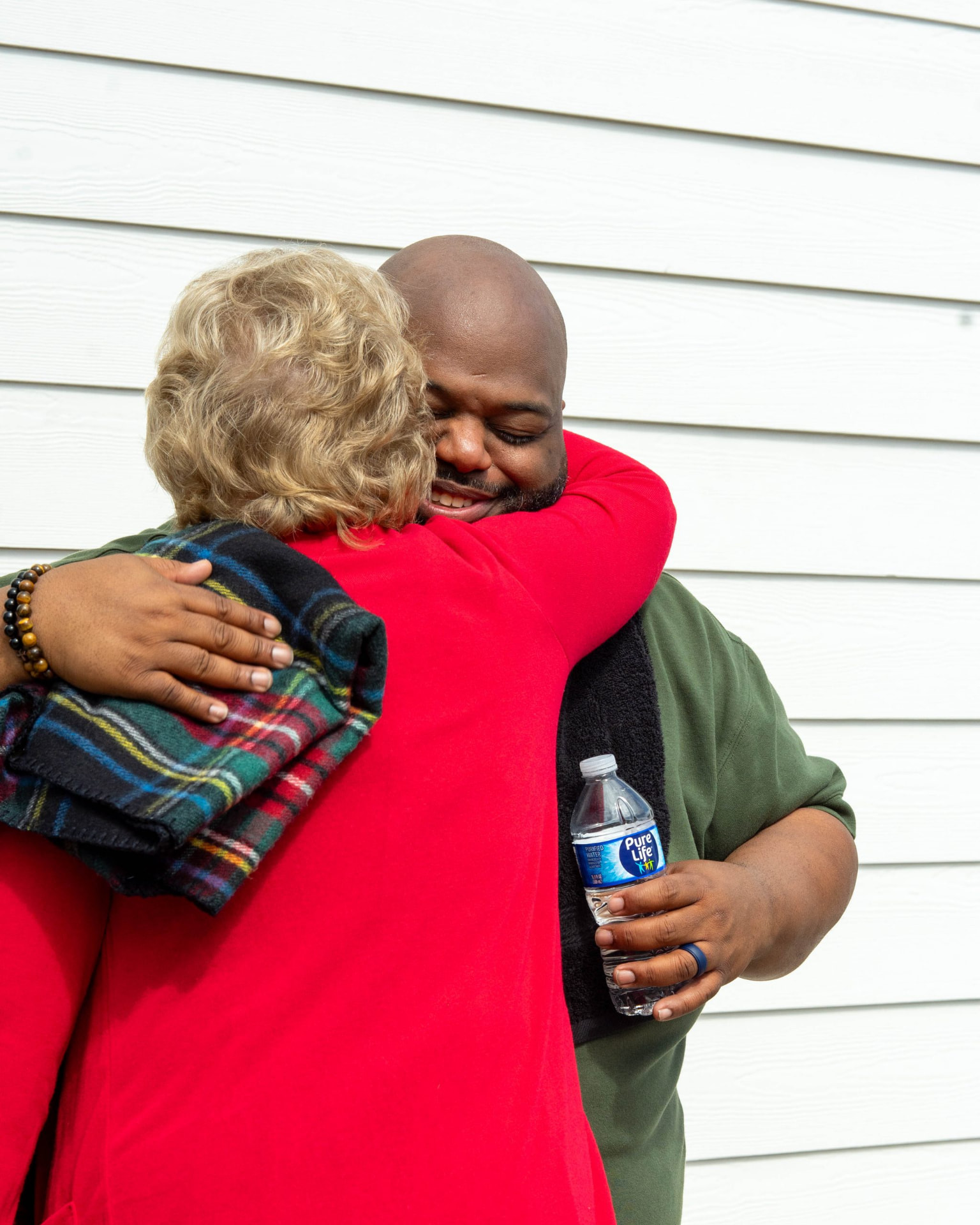 A woman in red embracing a smiling man holding a water bottle outside a building with white siding.