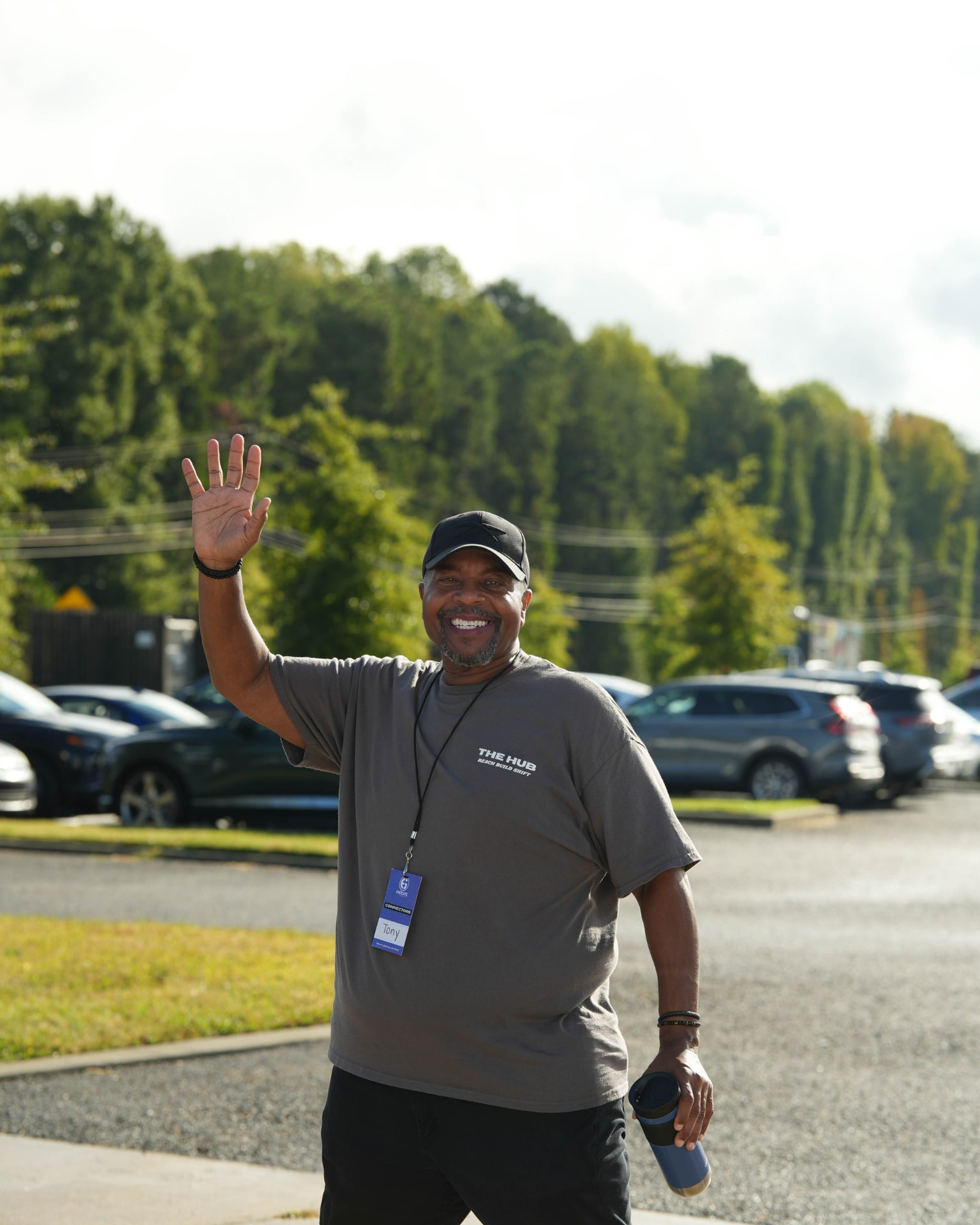 A smiling man in a cap and gray t-shirt waving while standing in a parking lot.
