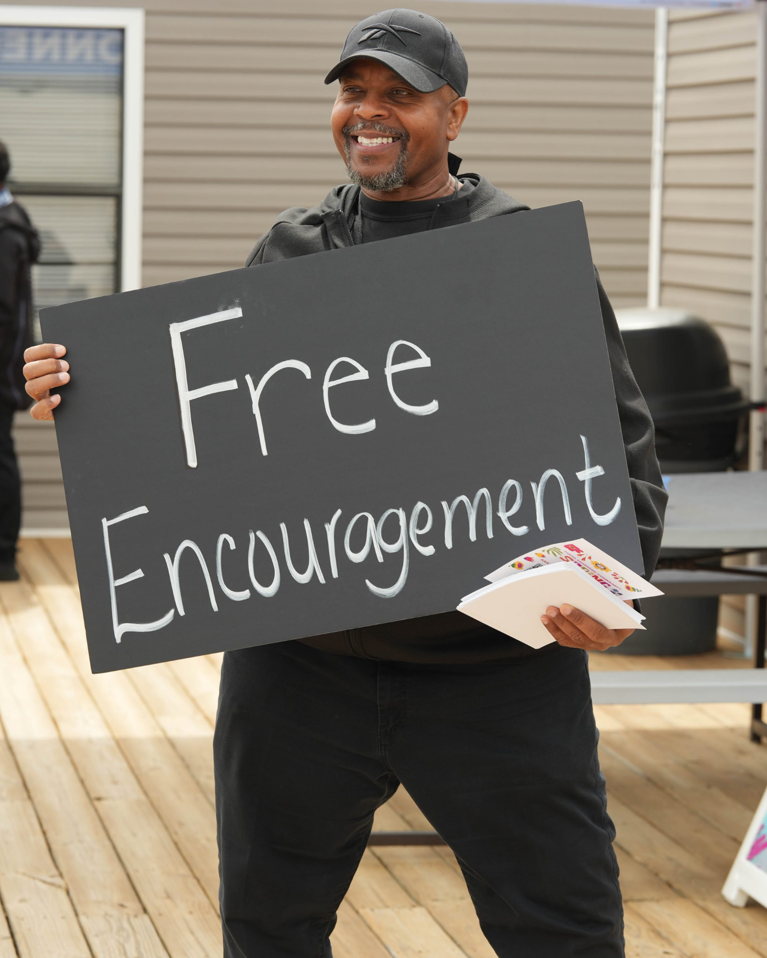 A smiling man in a cap and dark hoodie holding a large hand-lettered sign and a stack of cards.