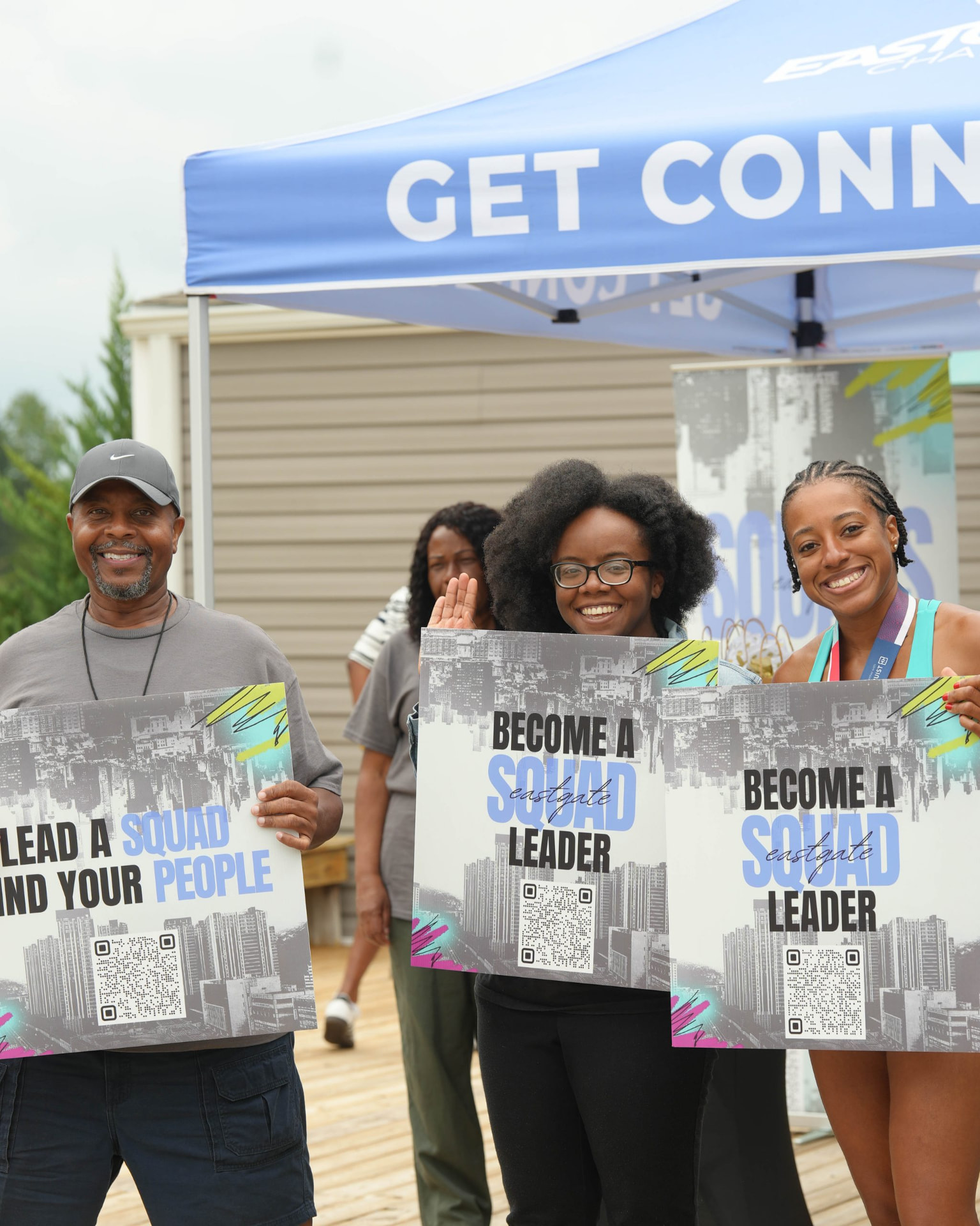 Three people smiling while holding large printed signs under a blue canopy.